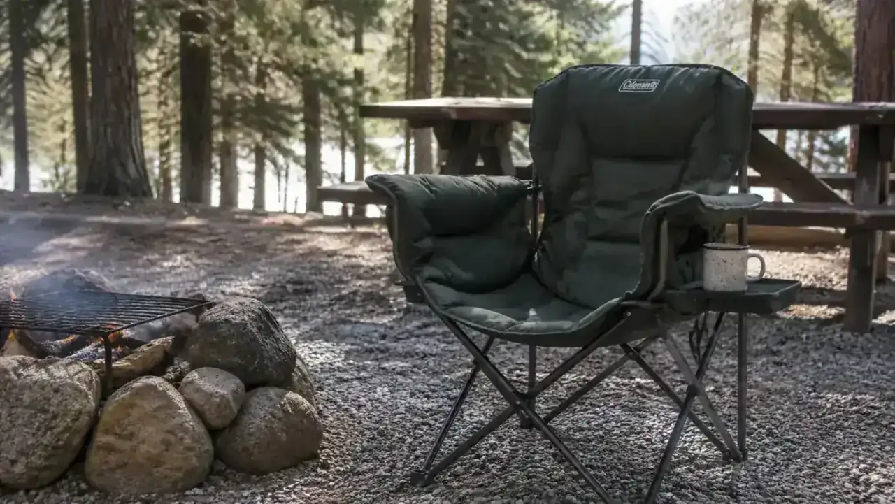 Coleman camping chair set up on a gravel campsite next to a fire ring, mid-morning light, empty coffee cup on the side table