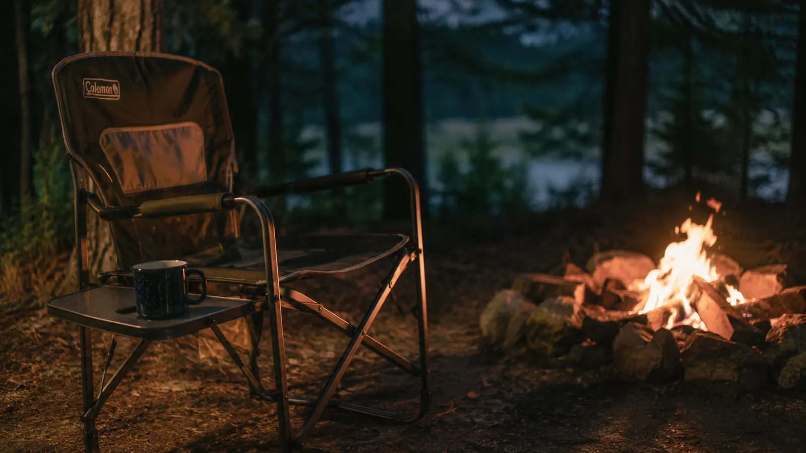 Coleman camping chair set up at a forested campsite in the evening, warm fire light, coffee mug on the side table