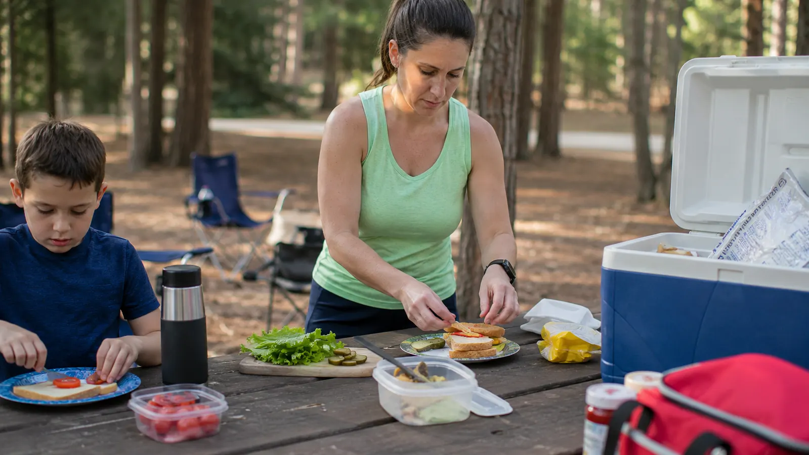 A well-packed hard-sided camping cooler sitting in the shade at a forested campsite, surrounded by food containers and a family preparing lunch