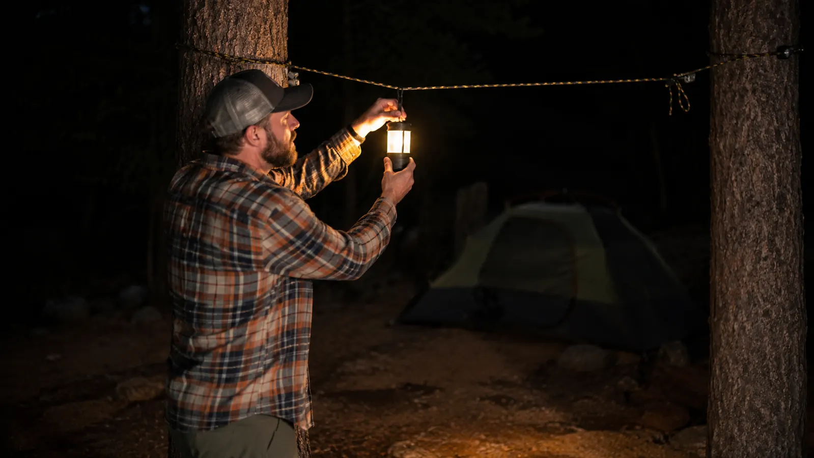 Person hanging a camping lantern from a carabiner clipped to a ridge line between two trees at night