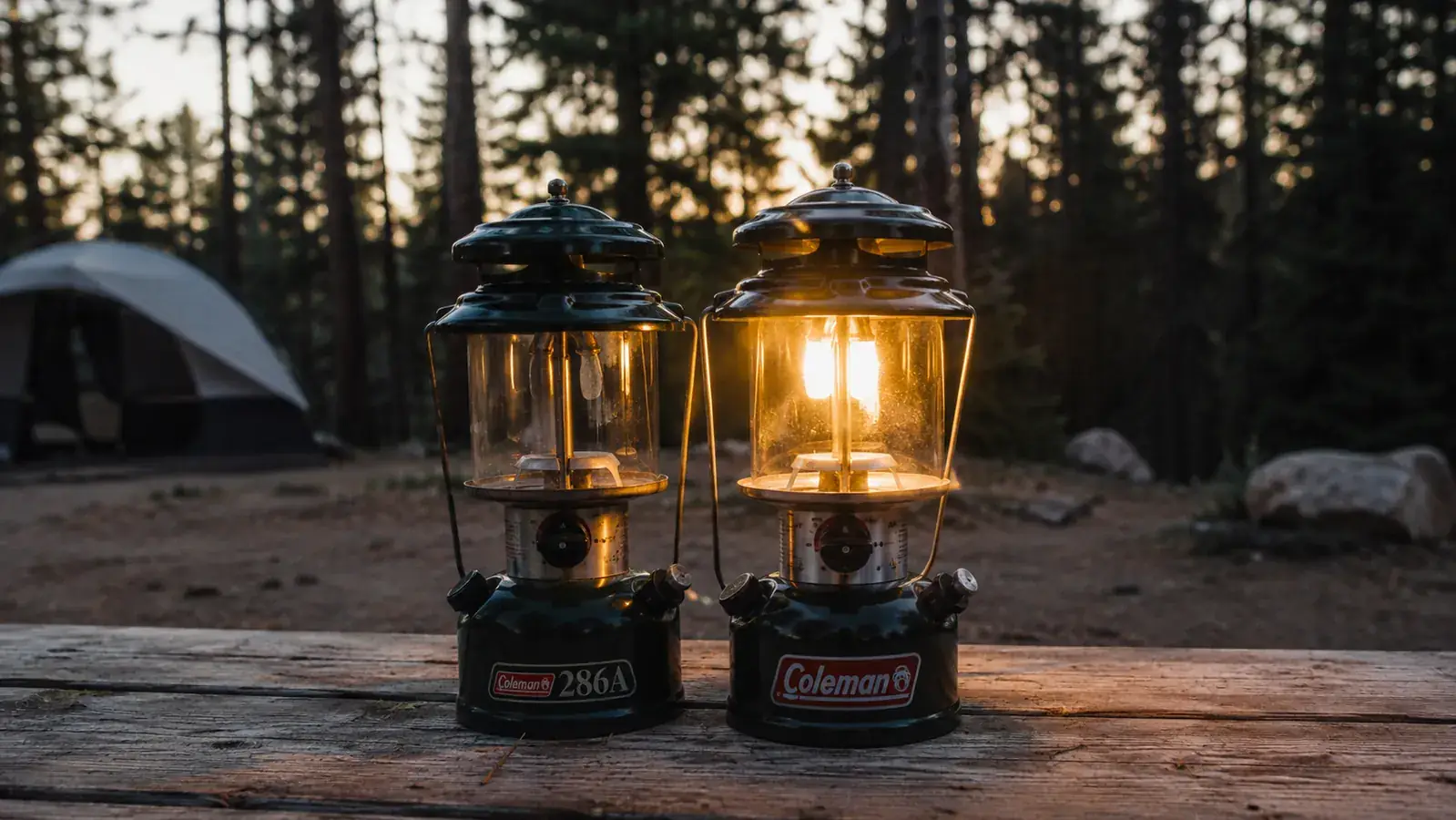 Two camping lanterns sitting on a picnic table at a forested campsite at dusk, one lit and casting warm light across the table